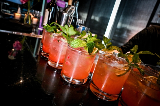 Half a dozen pink cocktails garnished with mint and lemon atop a black bar.