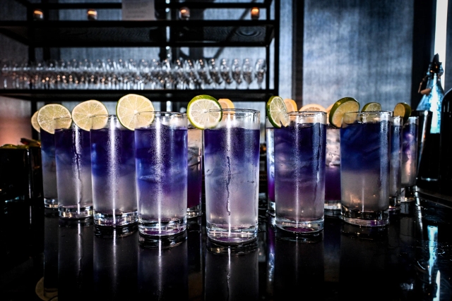 A dozen purple cocktails garnished with lime and lemon atop a black bar.