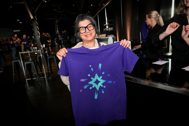 A woman with grey hair and black glasses smiling and holding up a purple Starlight t-shirt.