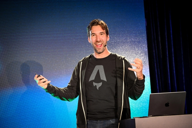 Fred K. Schott emoting enthusiastically with both hands outstretched, as he addresses the crowd onstage.