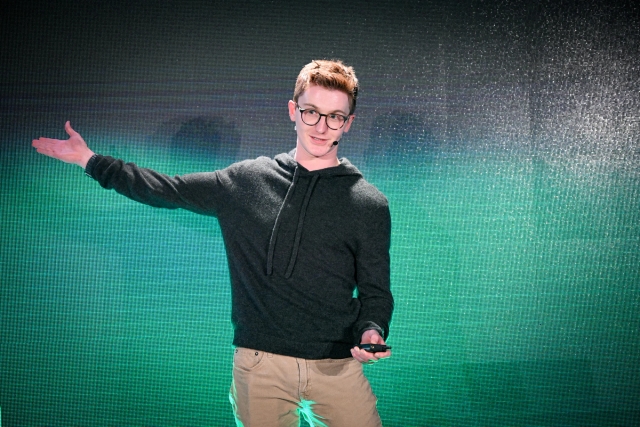 Ben Holmes on stage in front of a green and black LED screen. His right arm is outstretched, pointing to something off-camera.