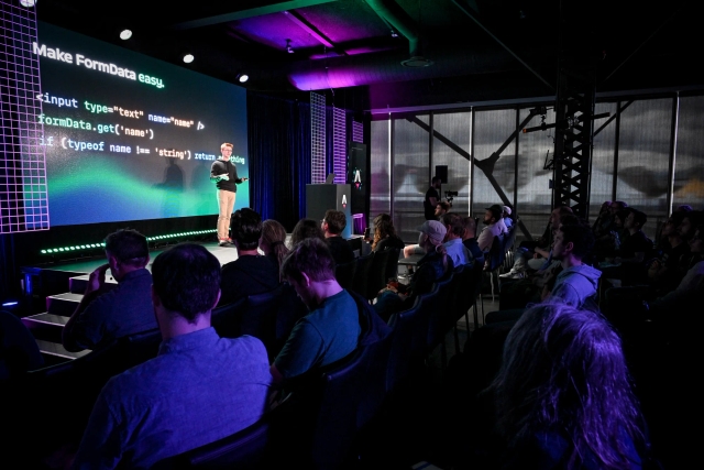 A shot from the side of the audience. Ben Holmes stands on stage in front of a code snippet depicting a form. The slide reads, "Make FormData easy."