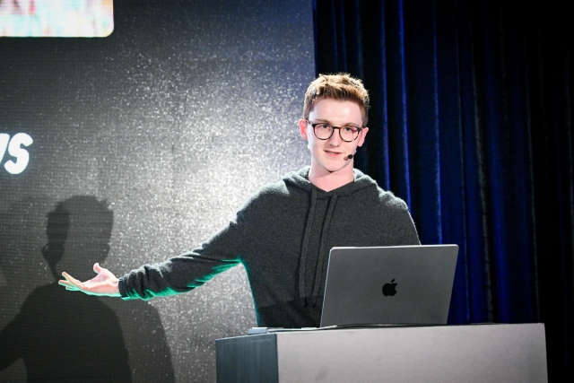 Ben Holmes on stage at the podium, gesturing to the LED screen off-camera.