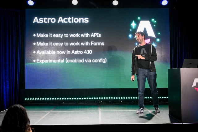 Fred K. Schott speaking on stage, pointing off-stage at the audience. Behind him is a summary of Astro Actions on the LED screen.