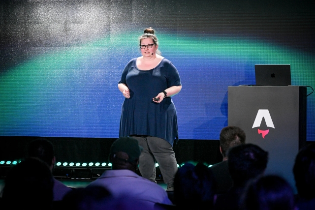 Sarah Rainsberger speaking on stage next to an Astro-branded podium, in front of a green and black LED screen.