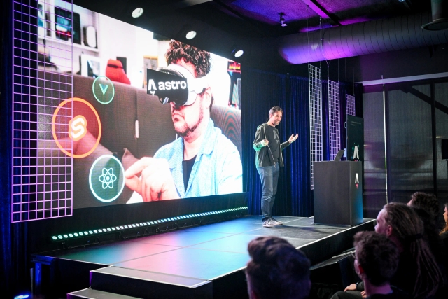 Side stage view of the front row and Fred K. Schott speaking on stage, making a joke about Astro using a meme of VR glasses.