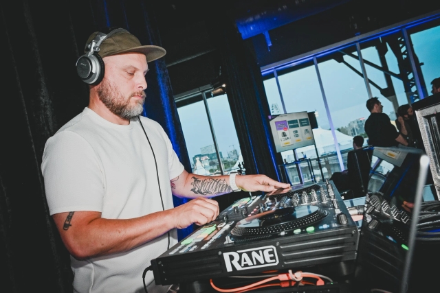 A DJ wears a cap and headphones while mixing at his booth.